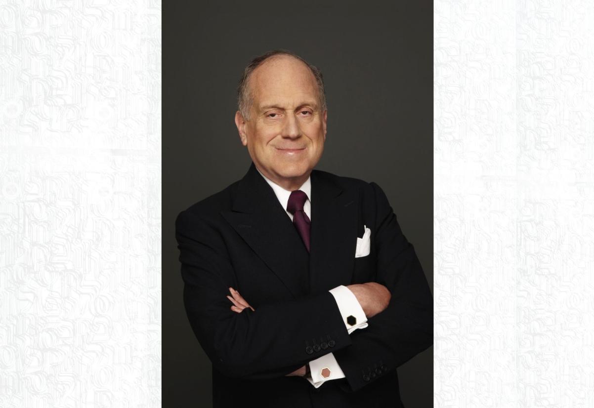 Ronald S. Lauder ubrany w czarny garnitur i elegancką białą koszulę stoi przodem do obiektywu z założonymi rękami. Uśmiecha się.