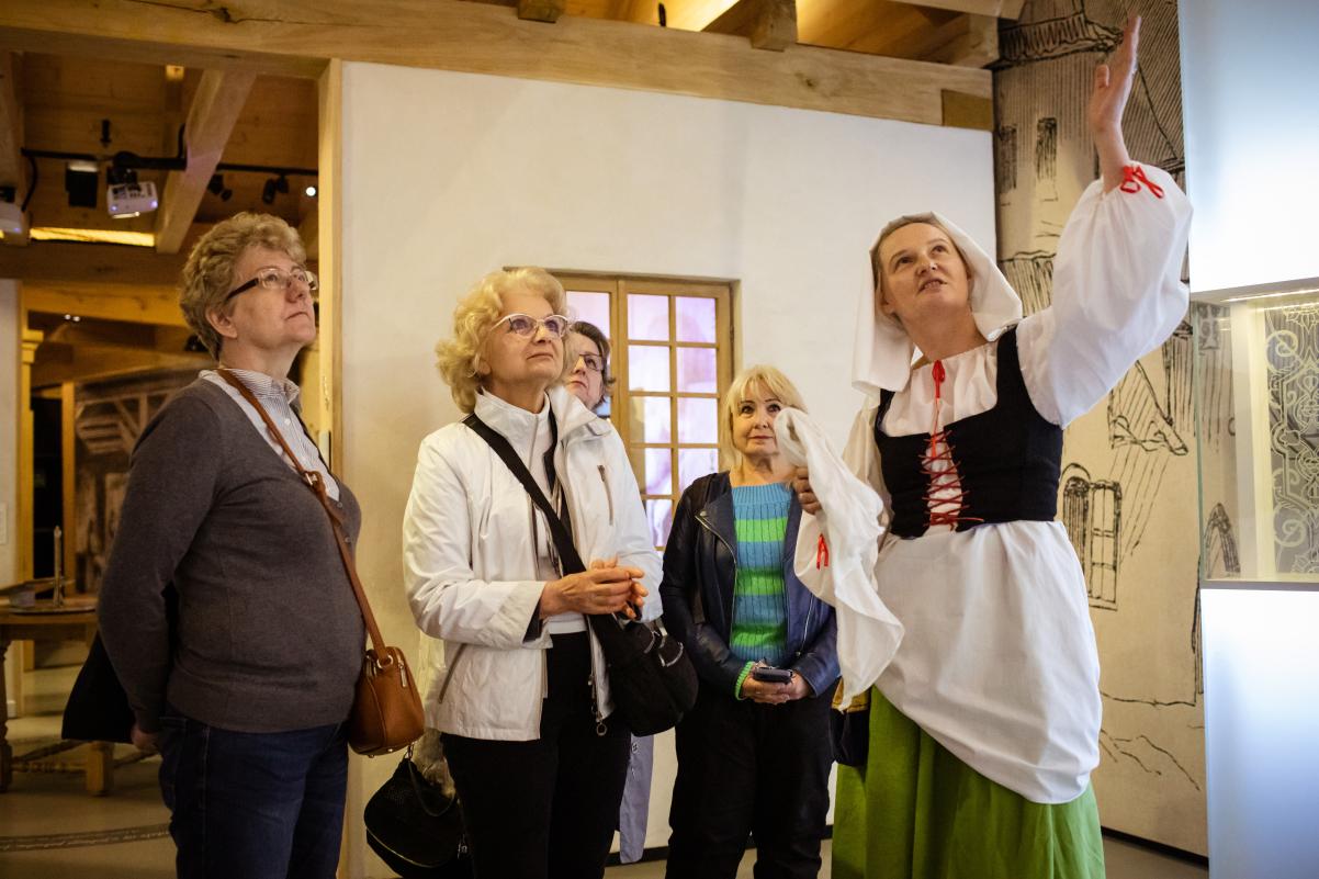 Grupa osób w wieku emerytalnym podczas zwiedzania wystawy stałej Muzeum POLIN. Przewodniczka ubrana jest w historyczny strój karczmarki.