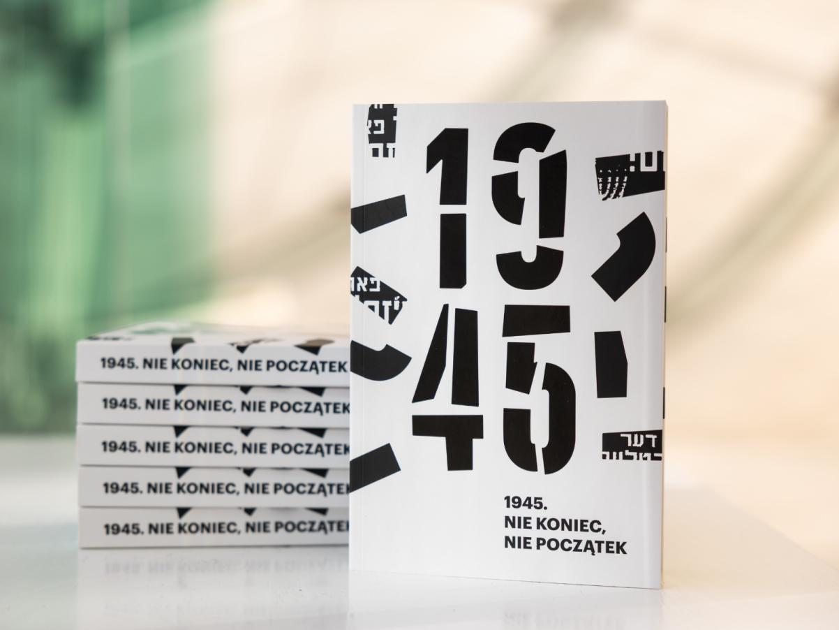Na stole stoi katalog wystawy "1945. Nie koniec nie początek", obok na stosie leży kilka egzemplarzy publikacji. W tle ściana w holu głównym Muzeum POLIN.