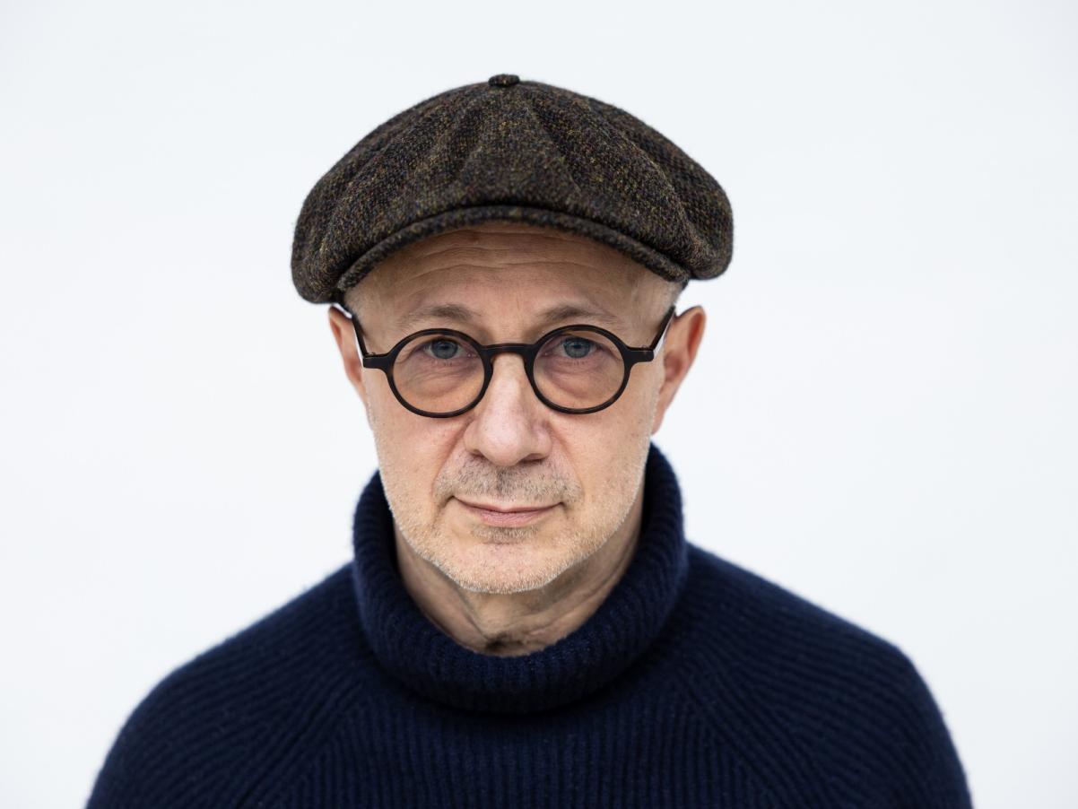 Mikołaj Grynberg - mężczyzna ubrany na czarno, na nosie ma okulary w ciemnych oprawkach, a na głowie szary kaszkiet.