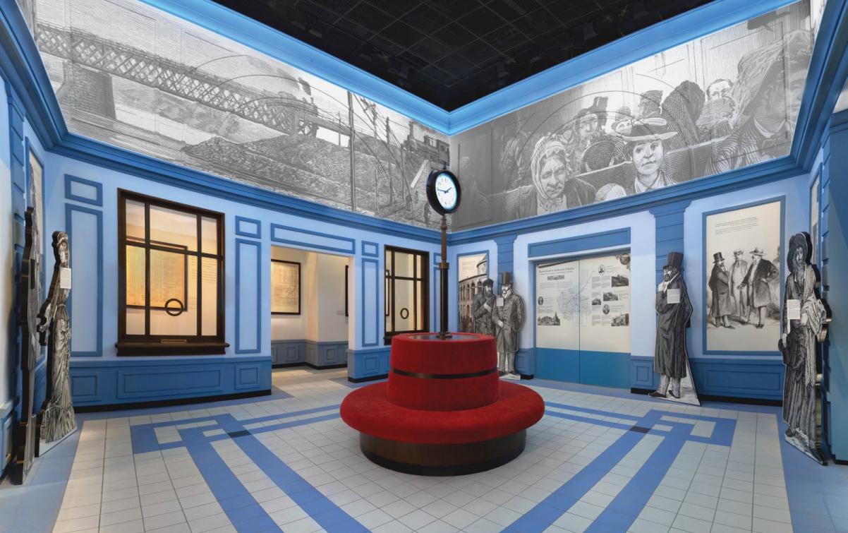 SALA DWORCA, ZREKONSTRUOWANA W PRZESTRZENI MUZEUM. NA ŚRODKU POMIESZCZENIA CZERWONE OKRĄGŁE SIEDZISKO, W KTÓRYM NA ŚRODKU TKWI NA RURZE WIELKI ZEGAR DWORCOWY.