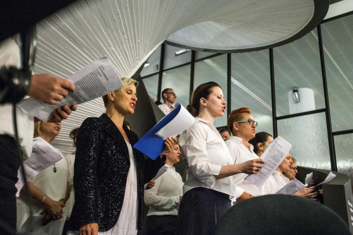 Chór POLIN - Czytanie wystawy Gdynia - Tel Awiw w Muzeum POLIN - na zdjęciu grupa dorosłych osób, ubranych galowo (białe koszule, czarne żakiety i garnitury. Śpiewają. Stoją pod krętymi schodami pomalowanymi na biało.