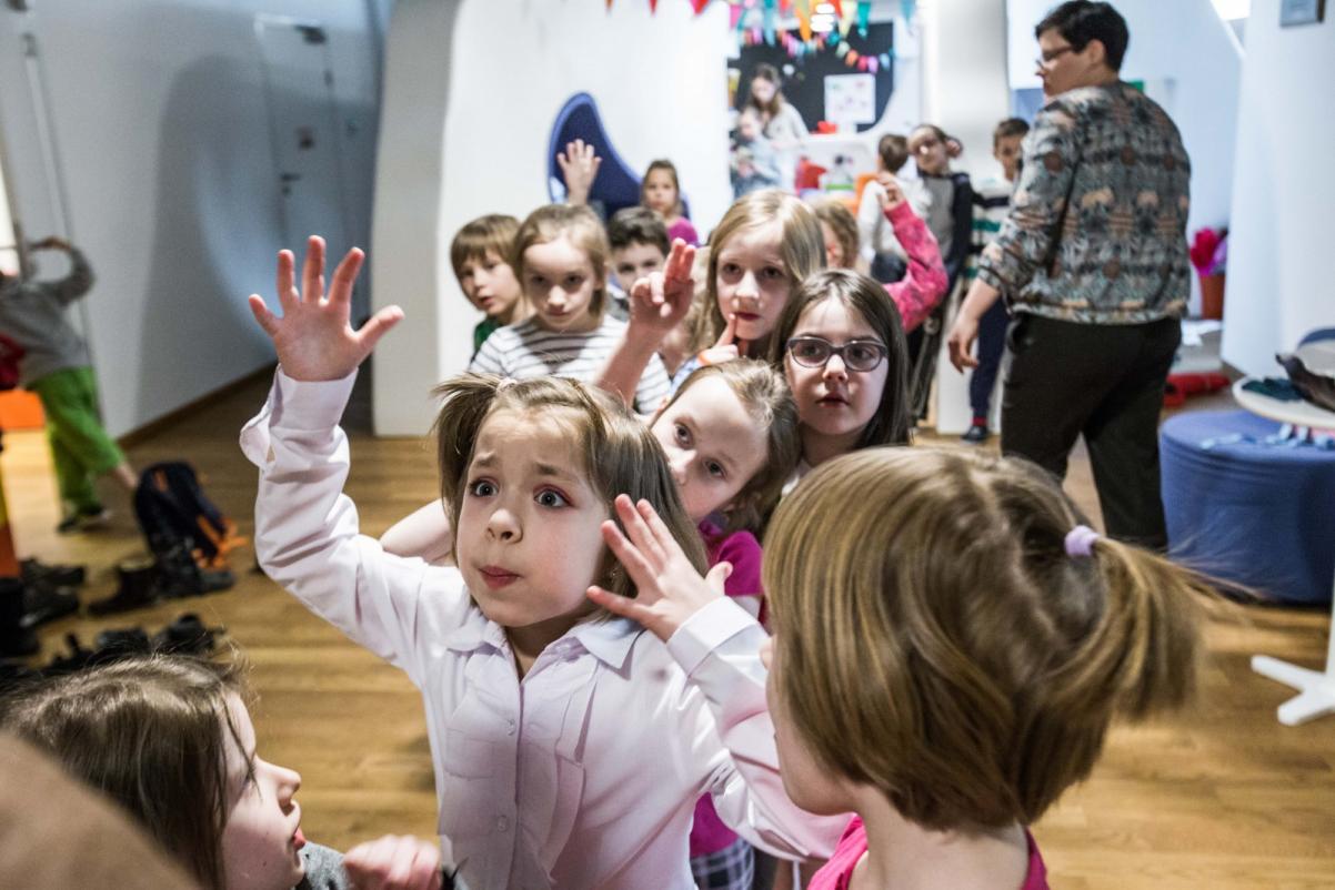 W sali z białymi ścianami stoi grupa dzieci. Na pierwszym planie dziewczynka w białej bluzce, która żywiołowo gestykuluje i coś tłumaczy.