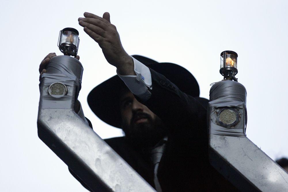 Żyd w kapeluszu unosi rękę do góry. Po jego obu stronach stoją kapliczki z małymi lampionami na górze.