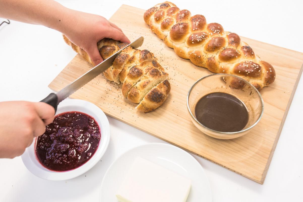 na desce kuchennej krojona nożem chałka, obok druga niepokrojona i dwie miski ze słodkimi dodatkami