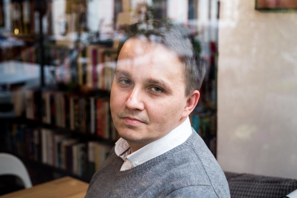 Nominowani do Nagrody POLIN 2019: Mirosław Tryczyk. Na zdjęciu Mirosław Tryczyk siedzi przy drewnianym stoliku patrzy przez ramię. W tle regał z książkami.