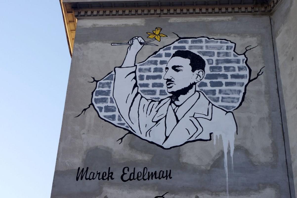 Mural przedstawiający młodego Marka Edelmana, ujęty z profilu, sylwetka od pasa w górę, w uniesionej prawej dłoni ściska żółtego żonkila. Mural wykonany przez D. Paczkowskiego na ścianie szkoły przy ul. Karmeliskiej w Warszawie - ilustracja warsztatów dla uczniów klas 7-8 szkół podstawowych w Muzeum POLIN
