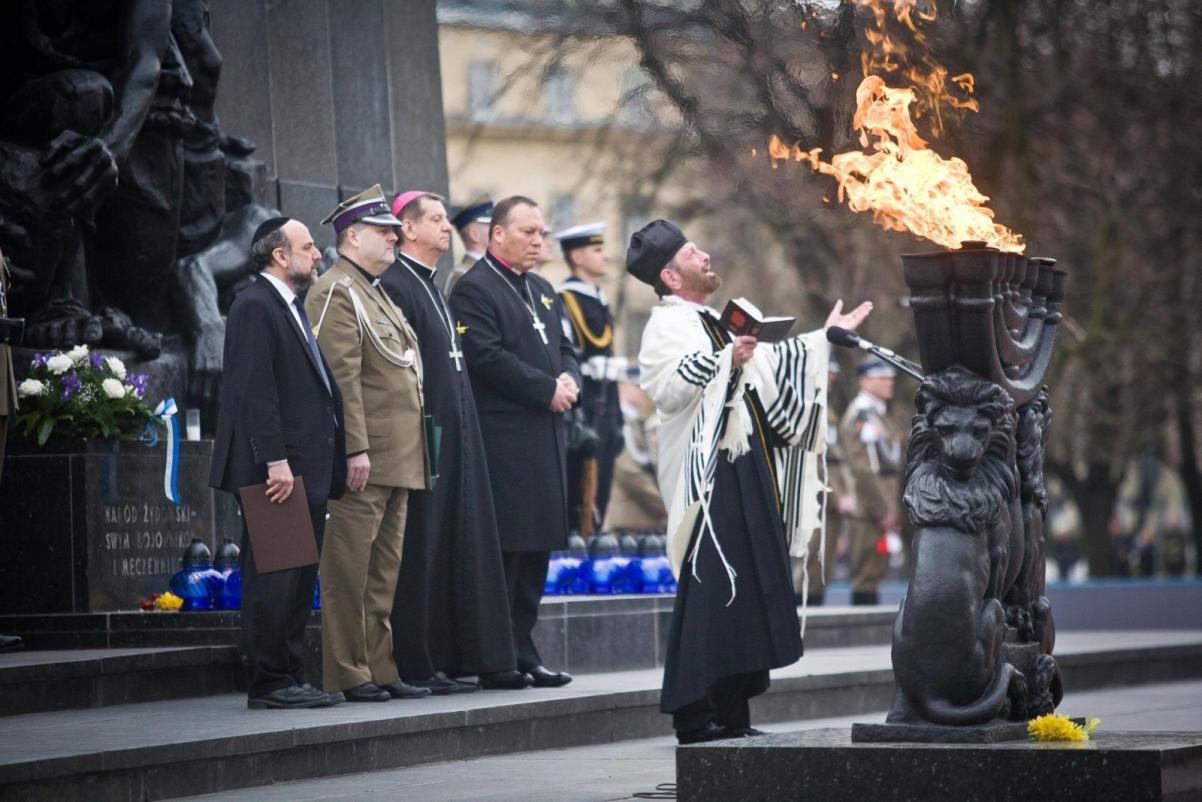 Grupa odświętnie ubranych osób stoi przed pomnikiem Bohaterów Getta. Pali się znicz.