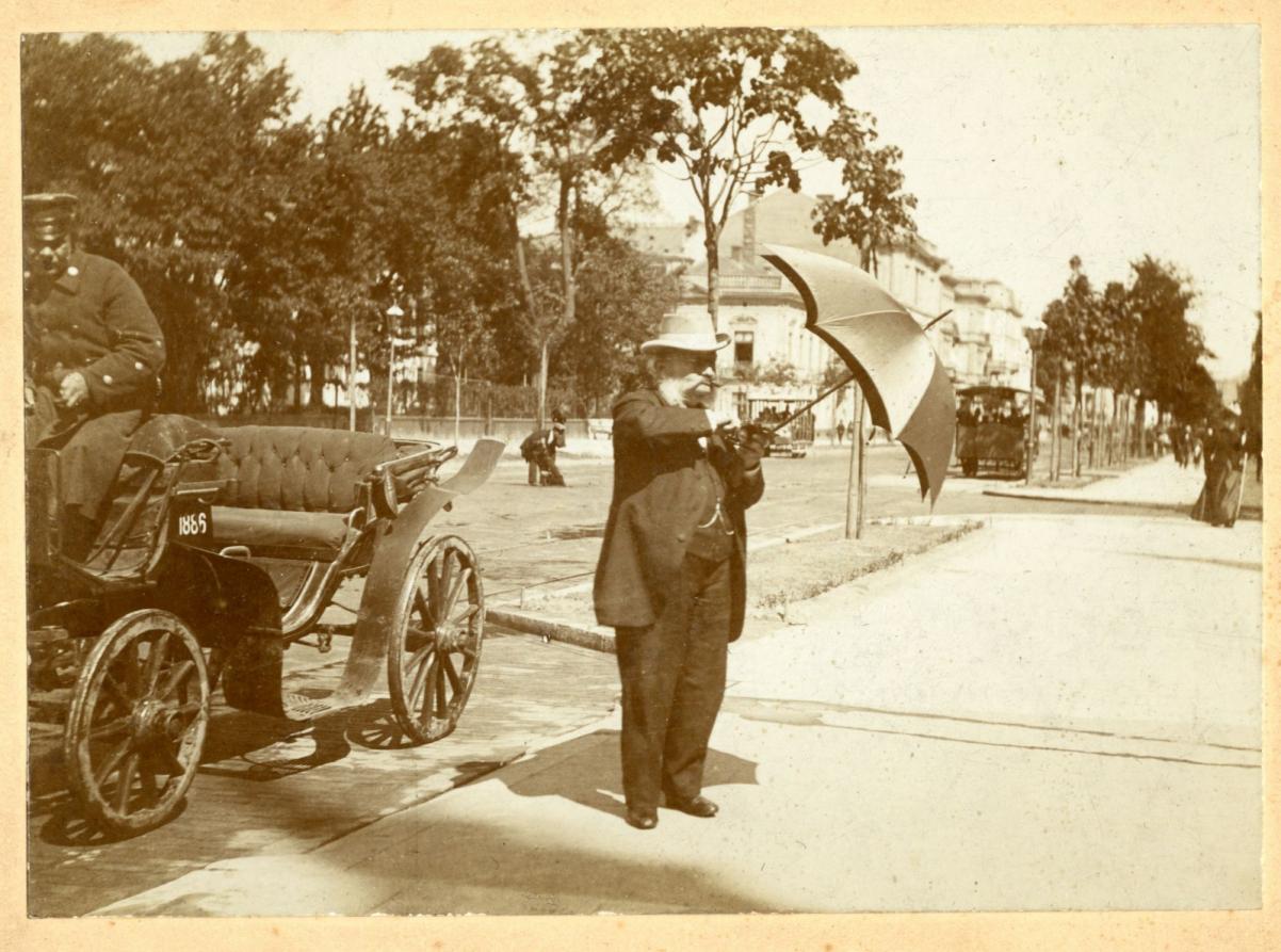 Mężczyzna stoi w parkowej alejce i otwiera parasol. Obok niego na wozie siedzi woźnica.