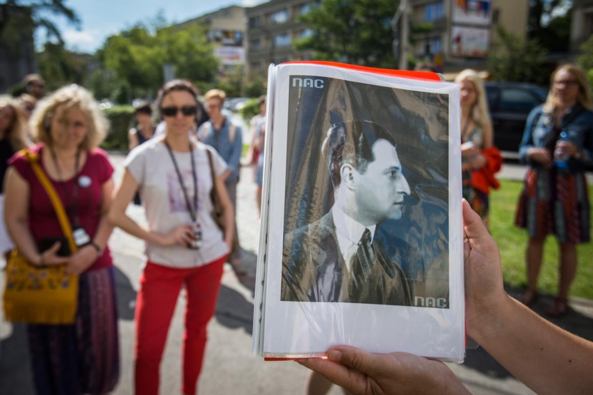 Ktoś trzyma czarno-biały portret Juliana Tuwima. W tle grupa uczestników spaceru miejskiego.