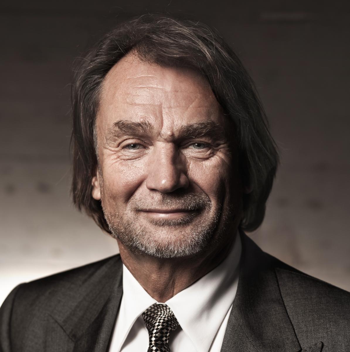 Jan Kulczyk. Mężczyzna w średnim wieku z ciemnymi włosami i lekkim zarostem. Ubrany jest w garnitur.