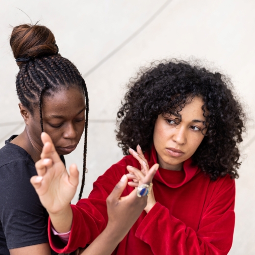 Two dark-skinned women are standing next to each other. They are holding their hands up, close to their faces.