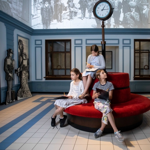 Trzy dziewczynki siedzą na kanapie w galerii "Wyzwania nowoczesności" na wystawie stałej w Muzeum POLIN. W dłoniach trzymają podkładki na kartki i ołówki. Three girls are sitting on a sofa in the Encounters with Modernity gallery at the Core Exhibition at the POLIN Museum. They are holding notepads and pencils in their hands.