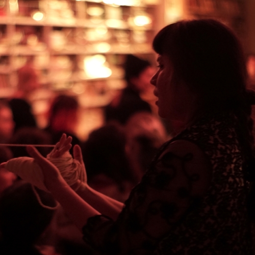The woman's hands are bound with white string. In the background, there is a group of other people.