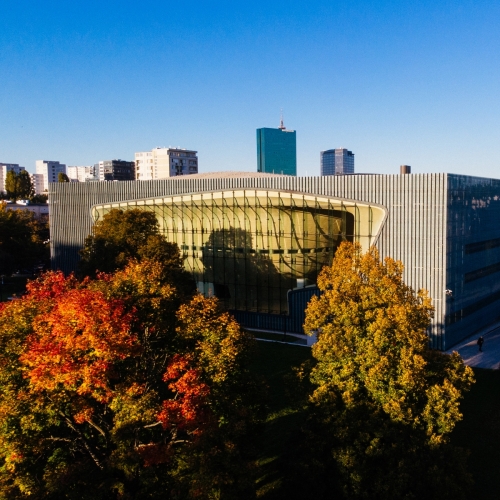 The POLIN Museum building surrounded by autumn trees. Skyscrapers in the background.