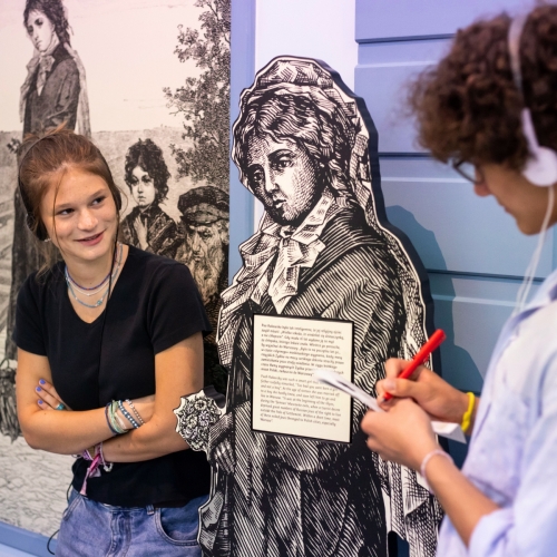 Wystawa stała w Muzeum POLIN. Dziewczyna patrzy na chłopaka, który coś notuje. Oboje są w słuchawkach.