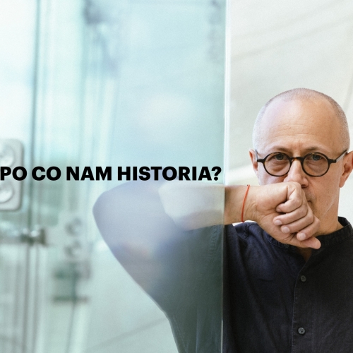 Mikołaj Grynberg oparty o szklaną ścianę w holu głównym Muzeum POLIN. Trzyma dłoń na brodzie.