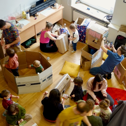 Dzieci i dorośli na zajęciach w klubie Kinderwelt. Niektóre dzieci siedzą w kartonowych domkach.