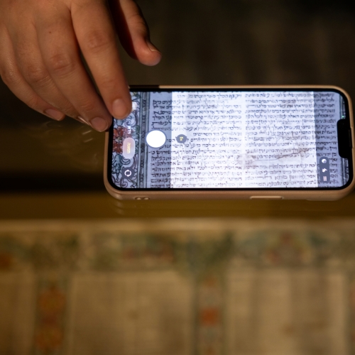Ktoś fotografuje telefonem pergamin zapisany w języku hebrajskim. Przykłada telefon do szyby gabloty.
