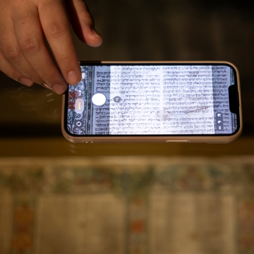Ktoś fotografuje telefonem pergamin zapisany w języku hebrajskim. Przykłada telefon do szyby gabloty.