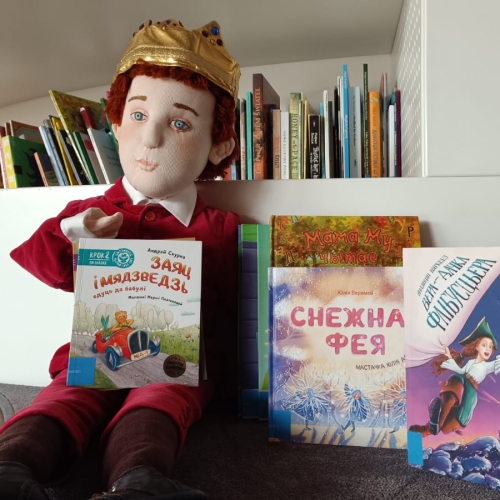 Лялька Кароль Мацюсь сядзіць каля палічкі з кнігамі ў атачэнні беларускіх кніг