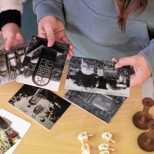 Dwie osoby trzymają czarno-białe fotografie podczas zajęć edukacyjnych w Muzeum POLIN. Na stole leżą też drejdle - drewniane bączki chanukowe.